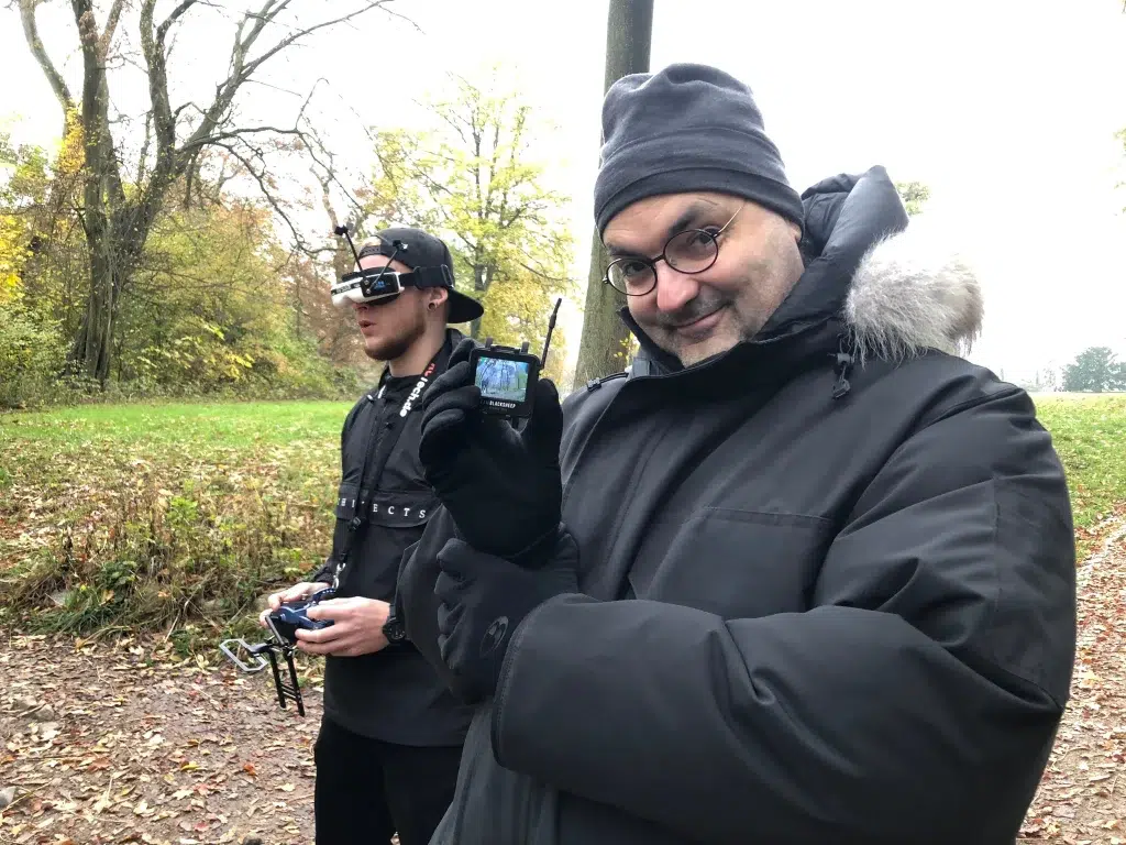 Zwei Personen stehen in einem Wald. Die Vordere Person blickt fröhlich in die Kamera und hält eine kleine Kamera in die Kamera. Die hintere Person trägt eine FPV-Brille und hält einen Controller in den Händen.