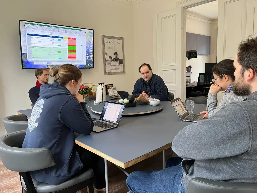 Eine Gruppe Personen sitzt an einem großen Tisch während einer Besprechung. Eine Person redet während die anderen zuhören. Im Hintergrund befindet sich ein Bildschirm auf dem detaillierte Informationen zum Drehplan stehen.