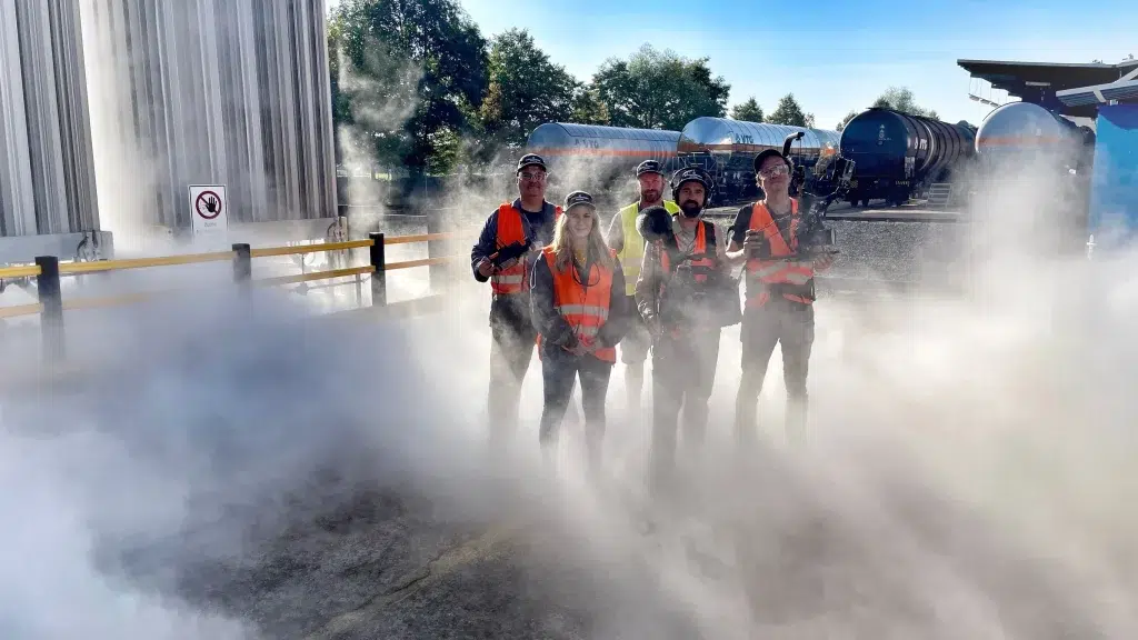 Ein Drehteam steht in Warnwesten in einem von Nebel umhüllten Industriegebiet. Sie halten Kameras und anderes technisches Equipment. Jim Hintergrund sind große Güterwaggons und ein Gebäude zu sehen.