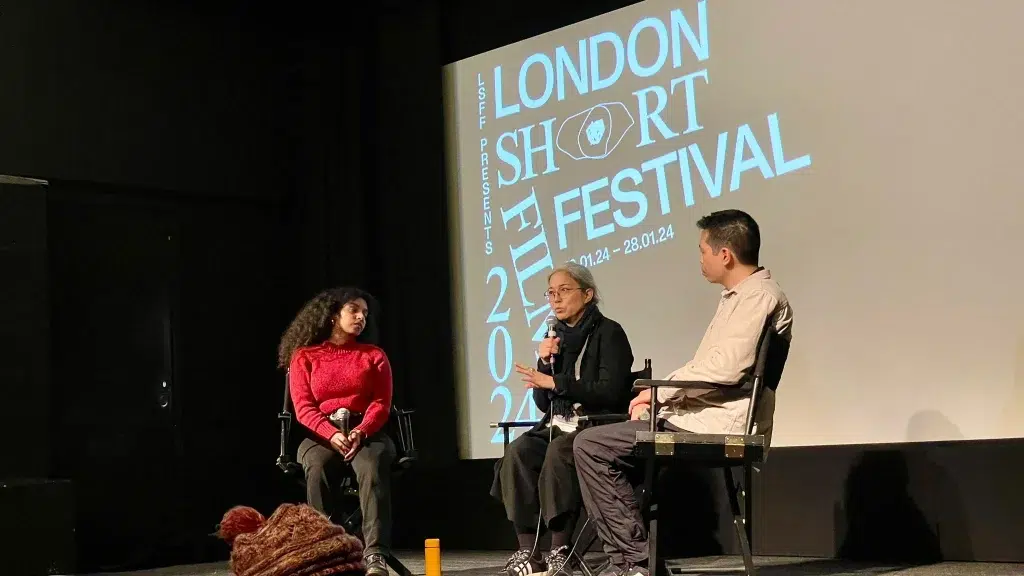 Drei Personen sitzen mit einem Mikrofon auf einer Bühne vor einer Leinwand mit dem Logo des London Short Film Festivals 2024. Marina Kem sitzt in der Mitte und spricht zum Publikum.