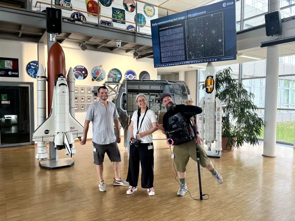 Eine Gruppe von drei Personen posiert fröhlich in einem Raum mit Modellen von Raumfahrzeugen. Links steht ein Modell des Space Shuttles Discovery und rechts ein Modell einer europäischen Rakete. Im Hintergrund sind ESA-Logos und ein großer Bildschirm mit wissenschaftlichen Bildern zu sehen.